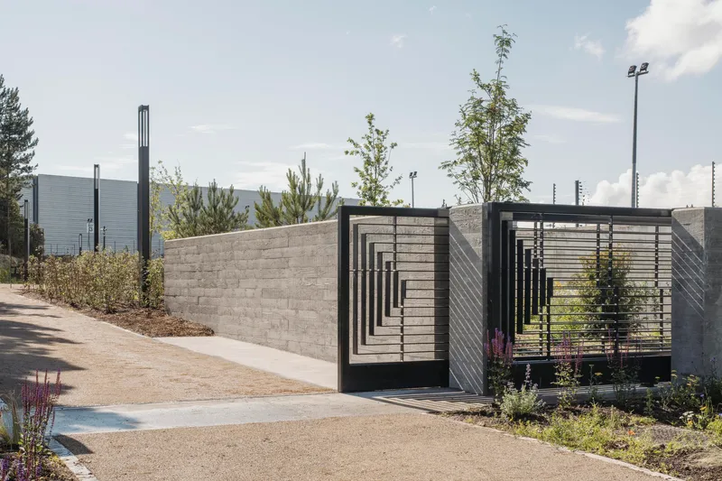 Concrete walls with black metal frame gateway and young trees along a paved pathway