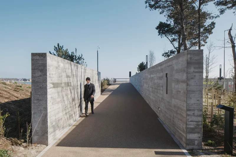 Board-formed concrete pathway flanked by tall walls with a figure walking and pines rising above