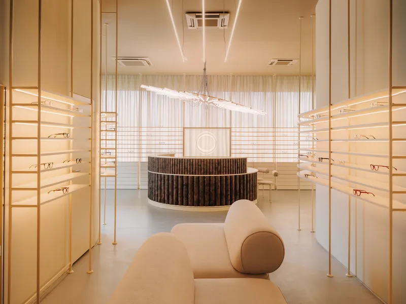 Interior view of an optical shop with backlit display shelves flanking a curved reception desk and linear ceiling fixture