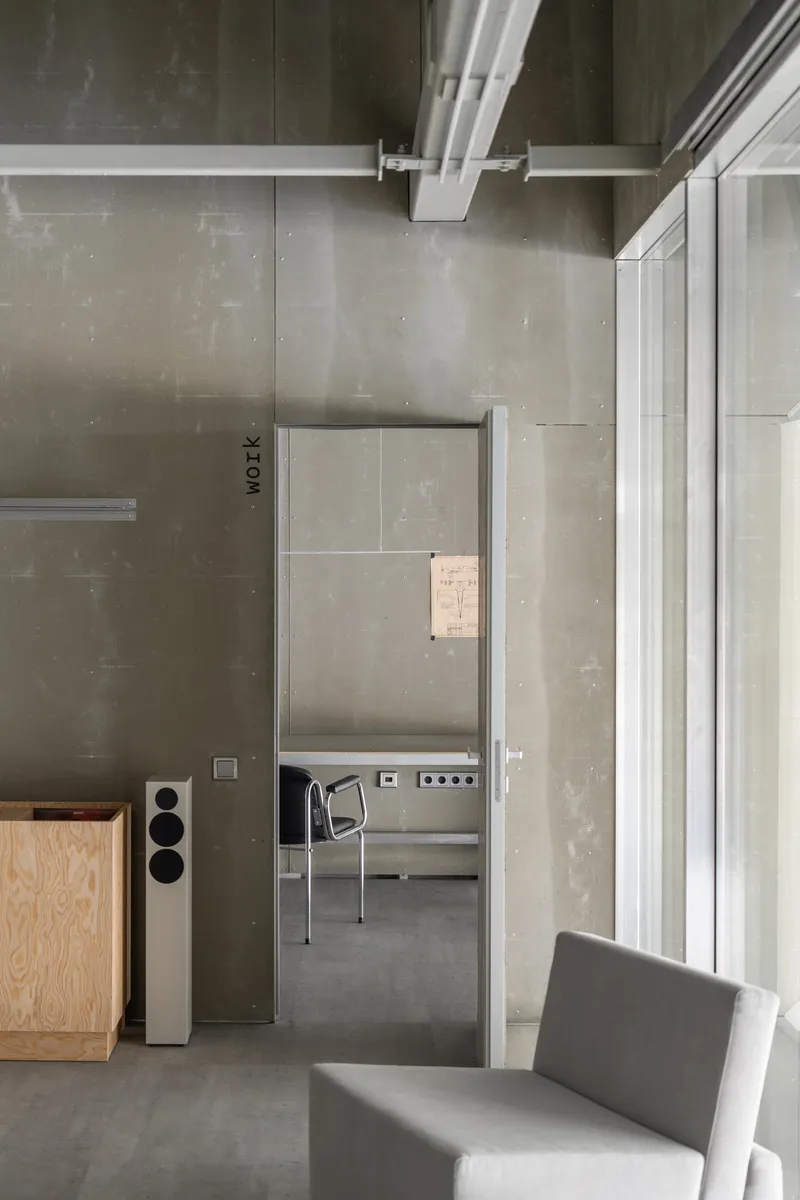 Doorway view through concrete walls with floor speaker and tubular steel chair visible beyond
