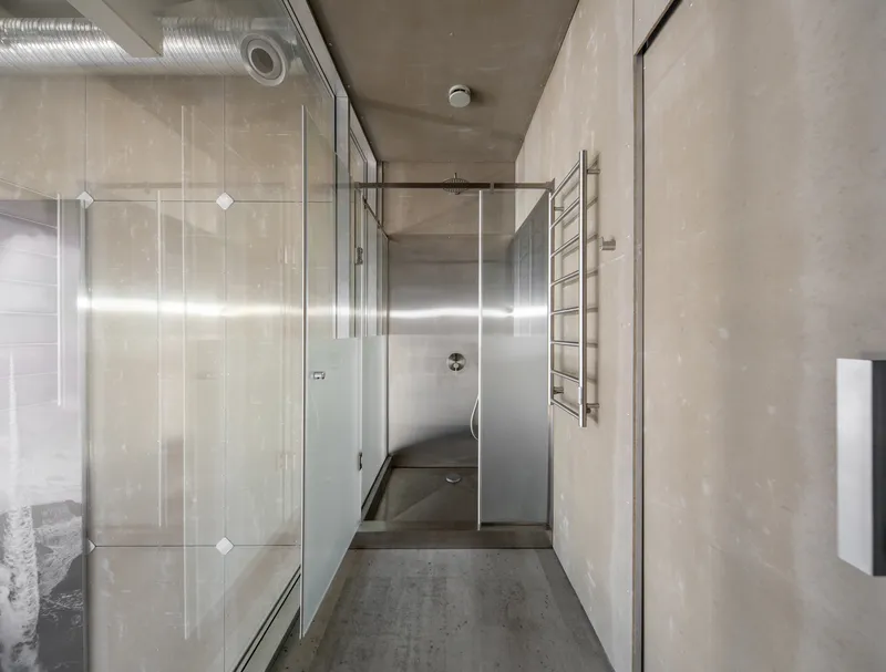 Corridor with glass shower enclosure, exposed concrete walls and ceiling, and industrial ducting overhead