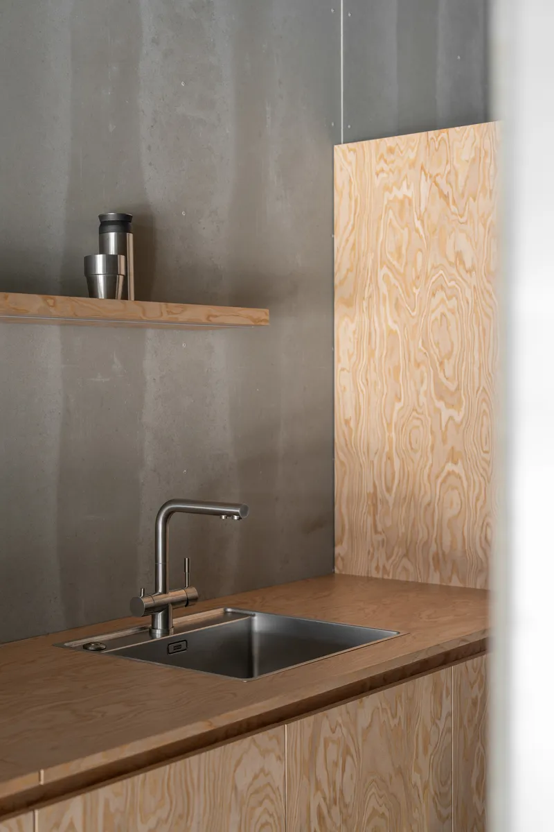 Kitchen corner with plywood cabinetry, stainless steel sink, and concrete wall with floating shelf