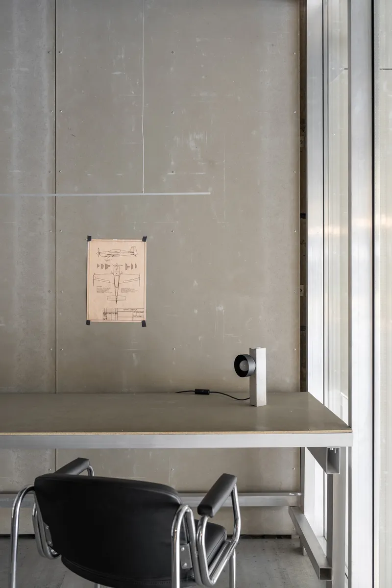 Workspace with concrete wall, window detail, suspended technical drawing, and black chair at desk