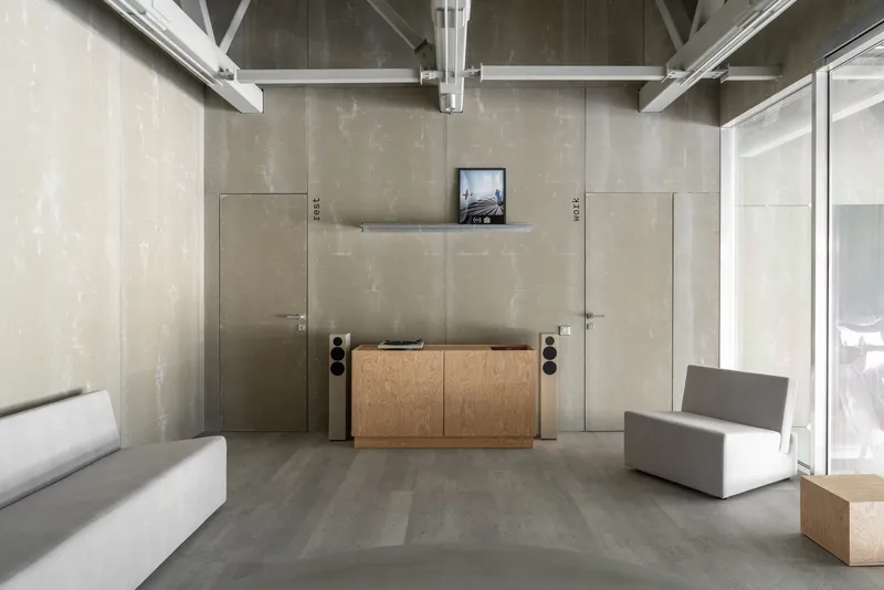 Living area with concrete panel walls, exposed white steel trusses, and plywood storage cabinet beneath a wall-mounted screen