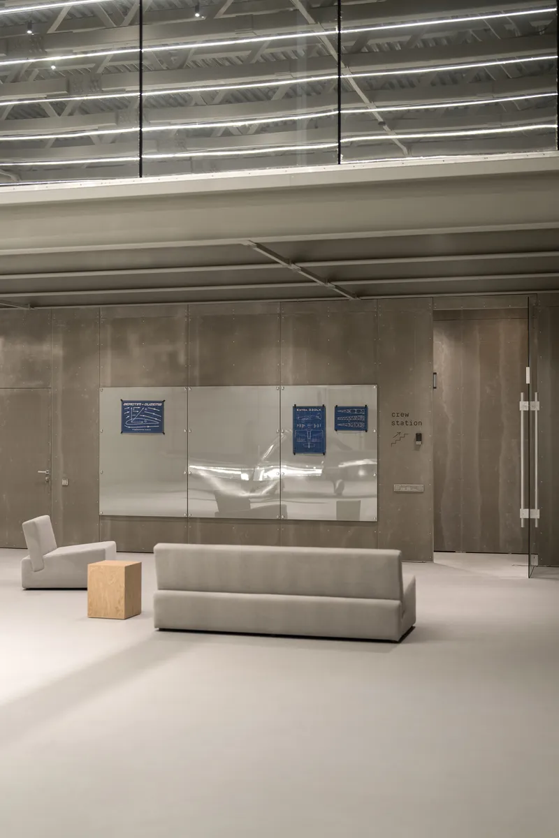 Interior lounge space with white board displaying project drawings mounted on concrete panels beneath exposed corrugated ceiling deck