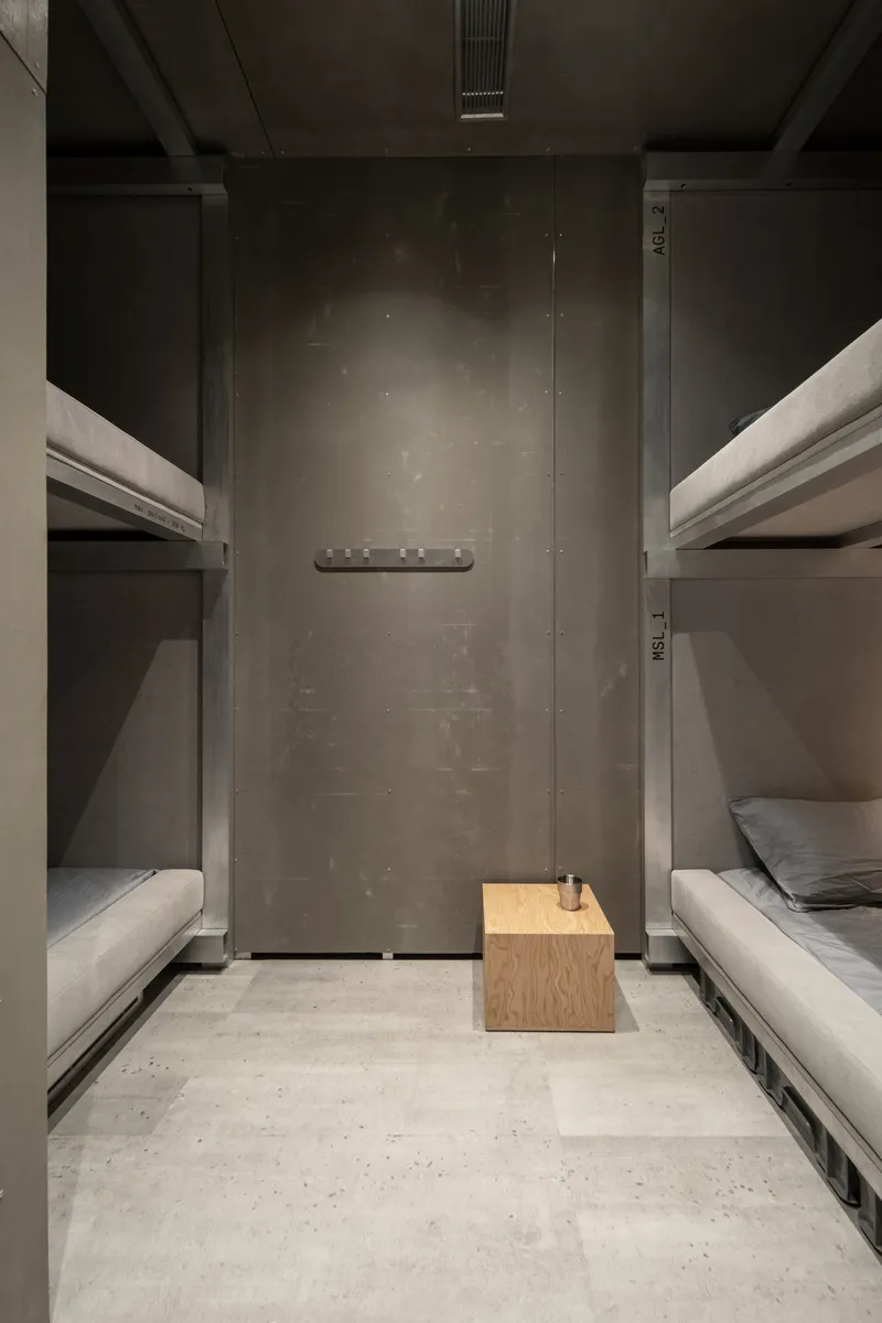 Narrow dormitory room with facing bunk beds, concrete walls, and a small wooden cube side table