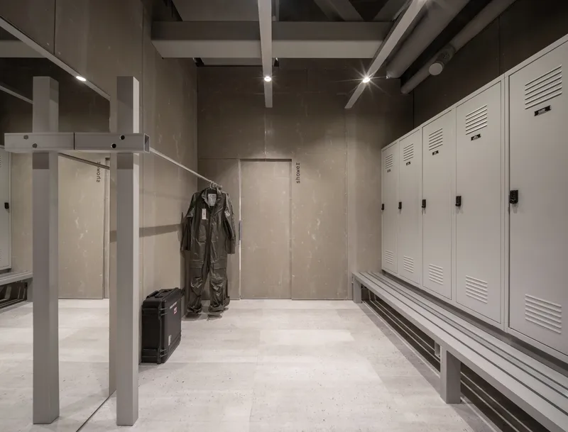 Locker room with white metal storage units, hung flight suit, and security scanner frame