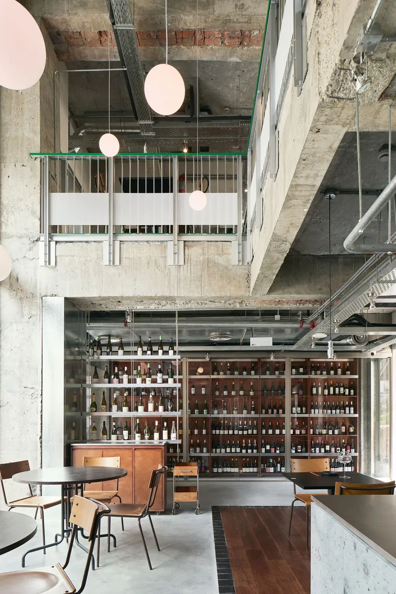 Double-height space with exposed concrete beams, suspended globe lights, and bottle-filled shelving behind a walnut bar