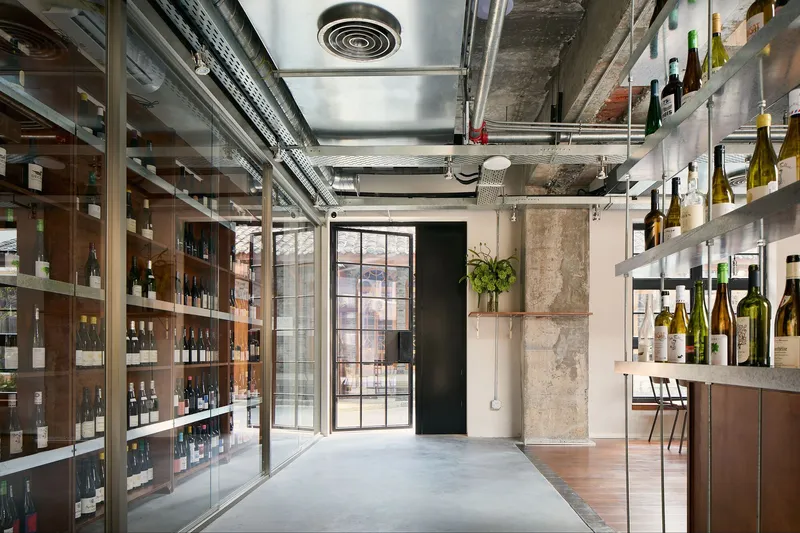 Entry corridor with glass-fronted wine shelving and gridded steel-framed doors opening to a courtyard