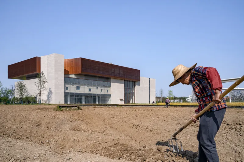 Farmer tilling bare earth in the foreground with the building's layered facade rising beyond young trees