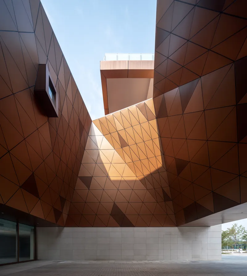 Upward view into a copper-clad triangulated courtyard with a cantilevered volume overhead in bright sunlight