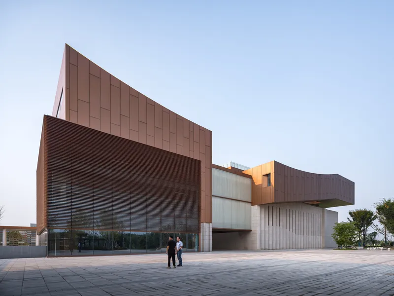 Plaza view of the stacked volumes clad in copper panels and vertical concrete fins at dusk