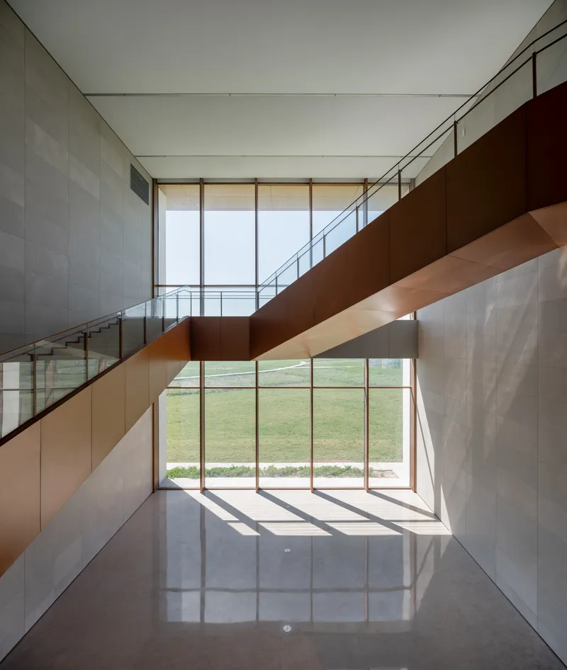 Double-height lobby with corten steel staircase and glazed curtain wall overlooking green lawn in daylight