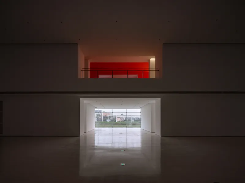 View through double-height void showing red-lit upper gallery and ground-level glazed entrance to landscape beyond