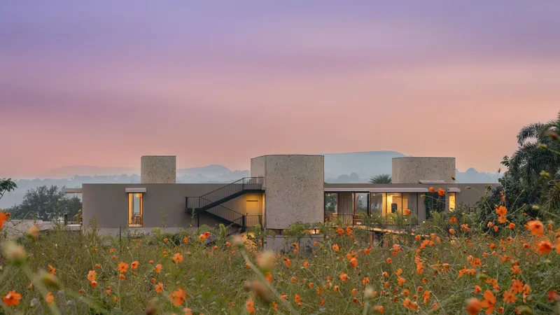 Stone-clad volumes with glazed openings and exterior stair framed by orange flowers under a pink sunset sky