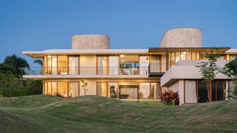 Two-story glazed facade with cylindrical stone towers and continuous balcony set on a sloping lawn at twilight