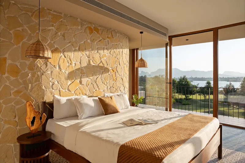 Morning light across the bedroom with stone wall and full-height glazing opening to lake views