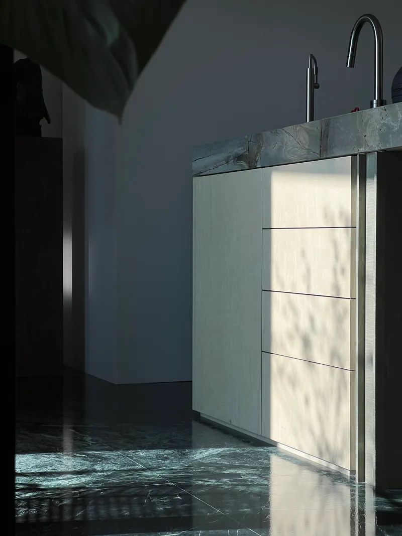 This architectural image showcases a modern kitchen design with a minimalist aesthetic. The composition highlights the interplay of light and shadow on the cabi