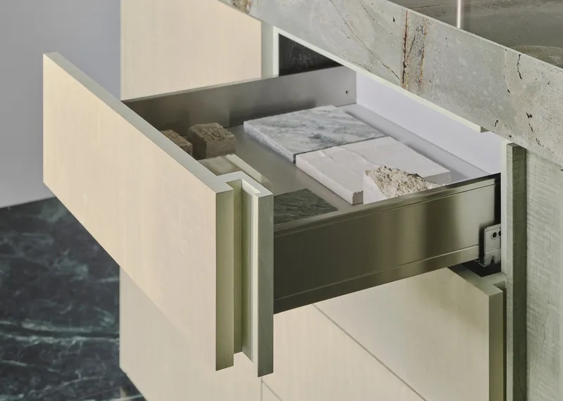 The image showcases a modern kitchen drawer, partially open to reveal an array of stone samples. The drawer features a light wood grain finish and is complement