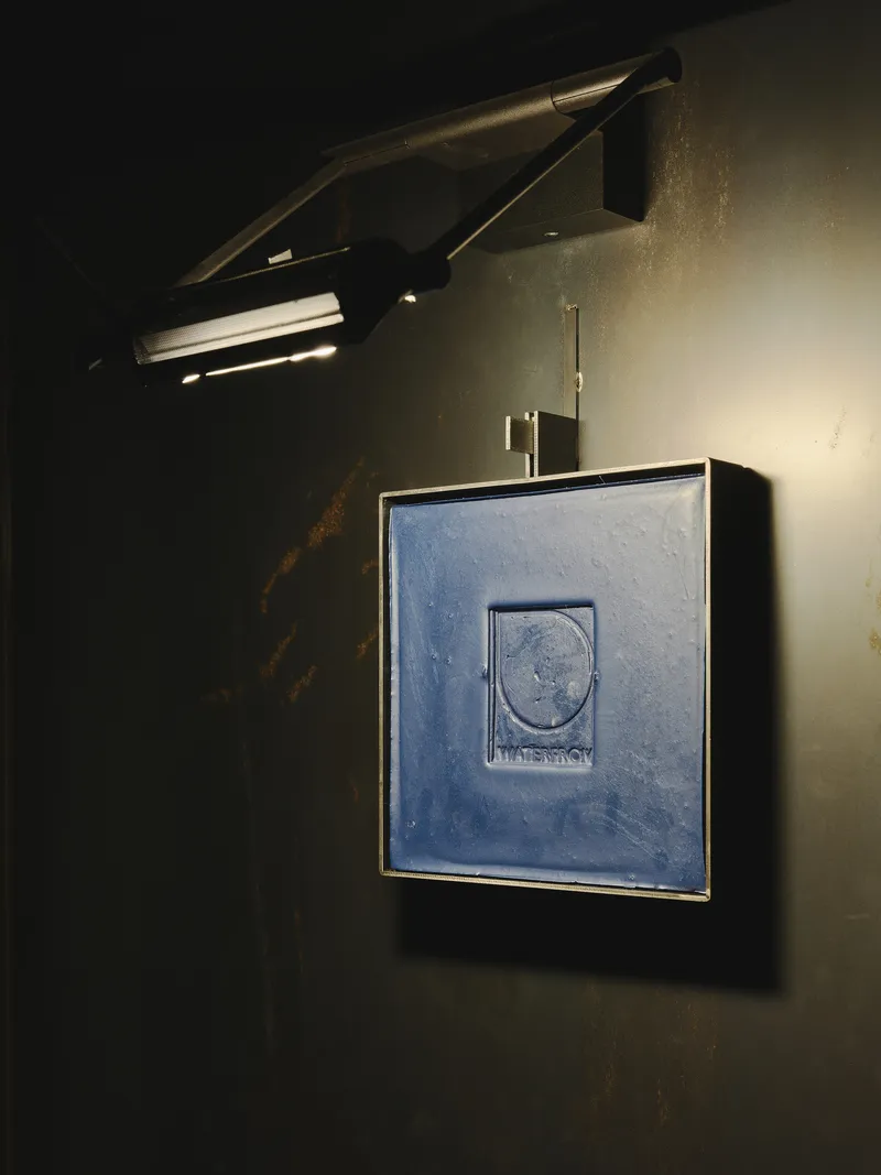 The image showcases a modern wall lamp illuminating a framed blue panel with the "WATERFROM" logo. The dark, textured wall and the interplay of light and shadow