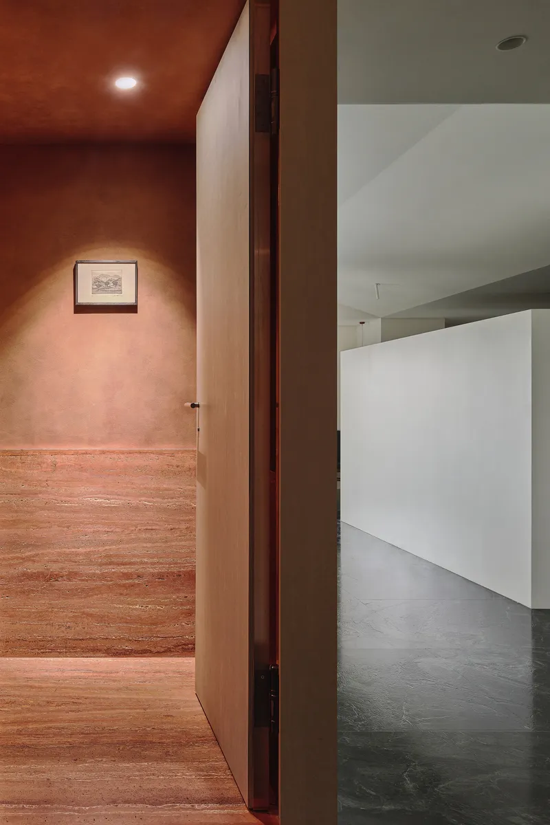 This architectural photograph showcases a doorway leading from a warm-toned, textured hallway into a minimalist, white-walled gallery space. The contrast betwee
