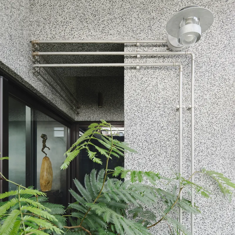 This architectural image showcases a modern building exterior with a textured terrazzo facade. A series of metal pipes and an outdoor light fixture are mounted