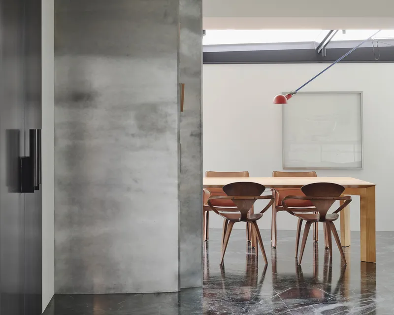 This architectural image showcases a modern dining room with a minimalist aesthetic. The space features a concrete wall with a sliding door, wooden chairs aroun