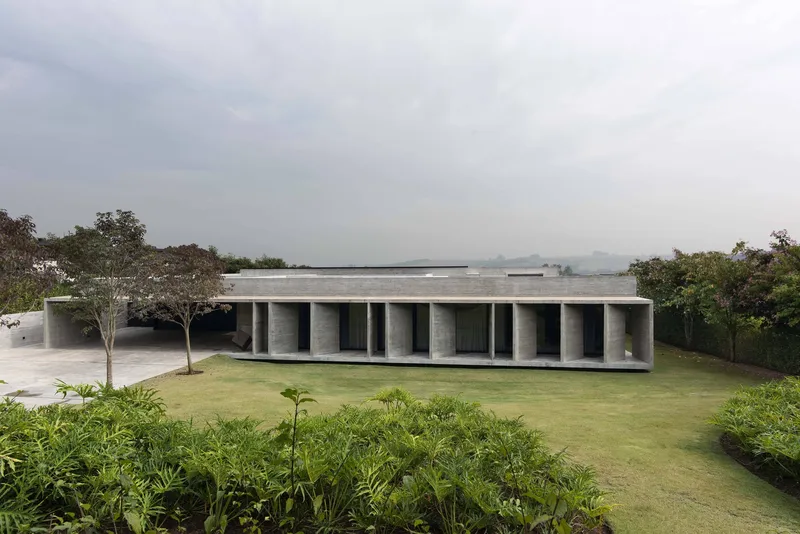 Long concrete pavilion with rhythmic columns set in lawn beneath trees and overcast sky
