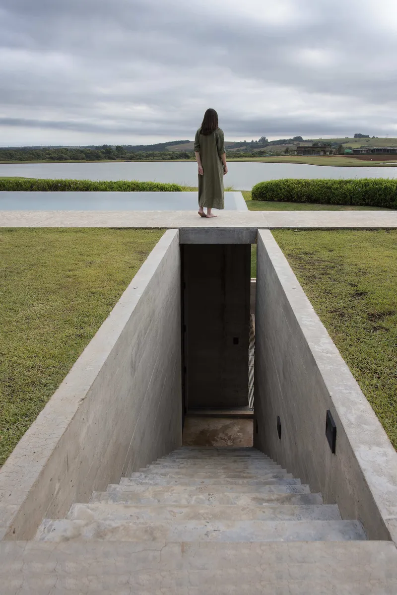Concrete stairwell descending from lawn with figure standing at terrace edge overlooking distant water