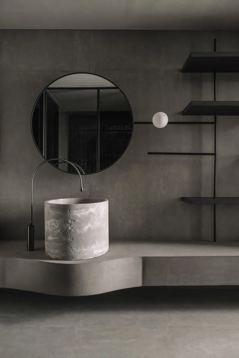 Bathroom vanity detail with cylindrical white stone basin and circular mirror against concrete wall