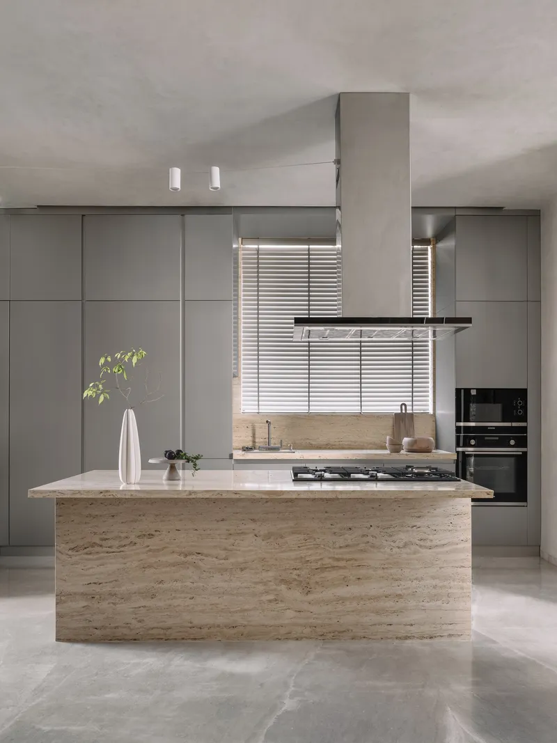 Kitchen island clad in travertine with grey cabinetry and timber blinds filtering window light