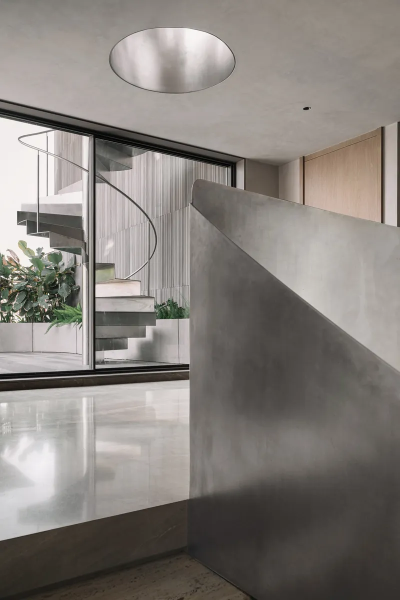 Curved polished concrete wall beside glazed opening revealing spiral stair and planted courtyard beyond