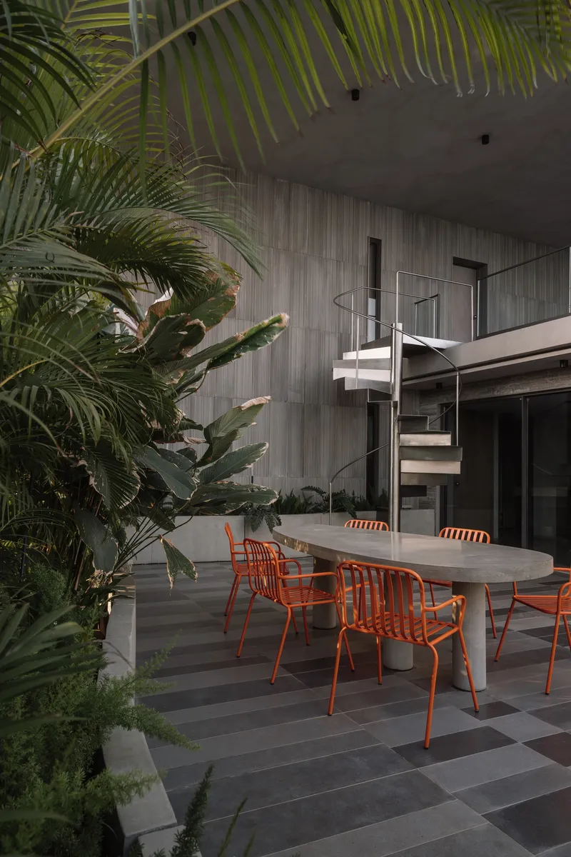 Outdoor dining terrace with concrete table and orange metal chairs surrounded by tropical palms at dusk