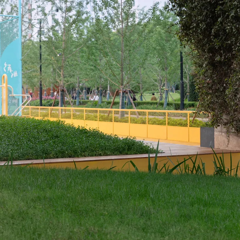 Yellow mesh bridge crossing a water feature with trimmed hedge and lawn beneath established trees