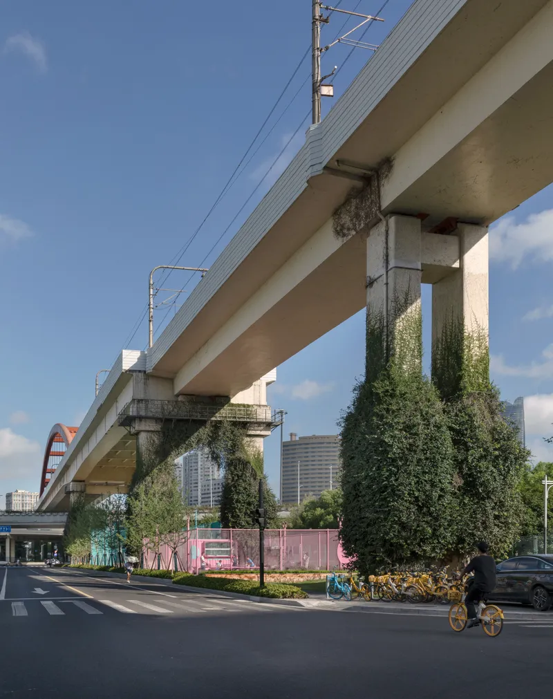 Vine-covered highway columns rising above pink-fenced sports facilities with a cyclist passing on the street