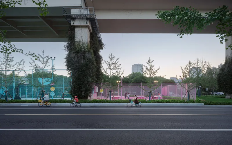 Street view of the pink mesh-fenced sports court beneath the elevated roadway with cyclists passing