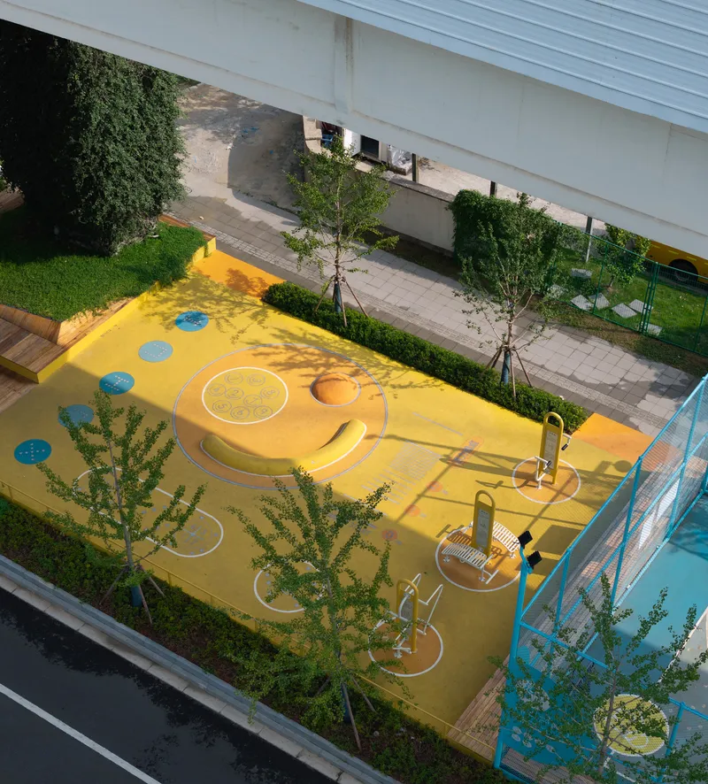 Overhead view of the yellow play area with circular elements and young trees casting shadows