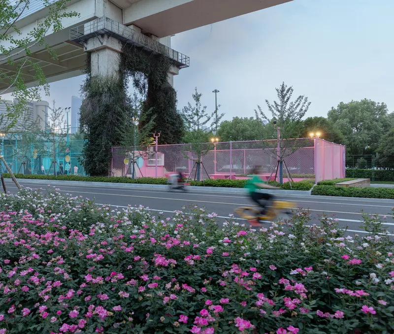 Pink mesh enclosure beneath a vine-covered viaduct pier with flowering beds and passing cyclists