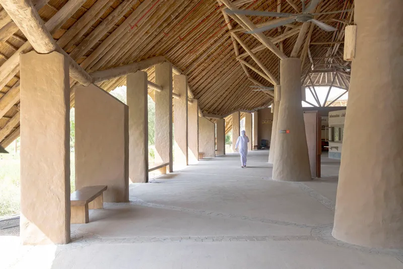 Covered walkway with plastered earthen walls and exposed timber roof structure as figure walks through