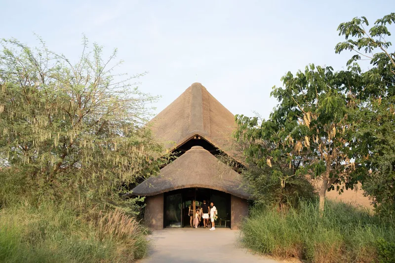 Entry pavilion with layered thatched roofs and arched opening as visitors gather at threshold