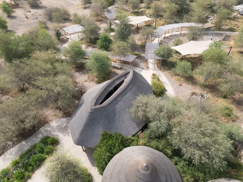 Drone view of crescent-shaped thatched roof nestled among scattered trees and pathways