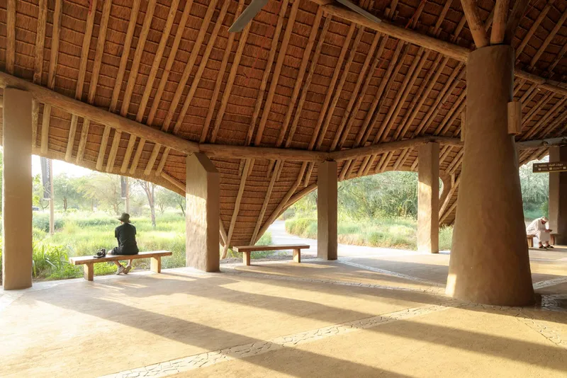 Open-air pavilion interior with exposed timber rafters and thatched ceiling sheltering benches and a seated figure