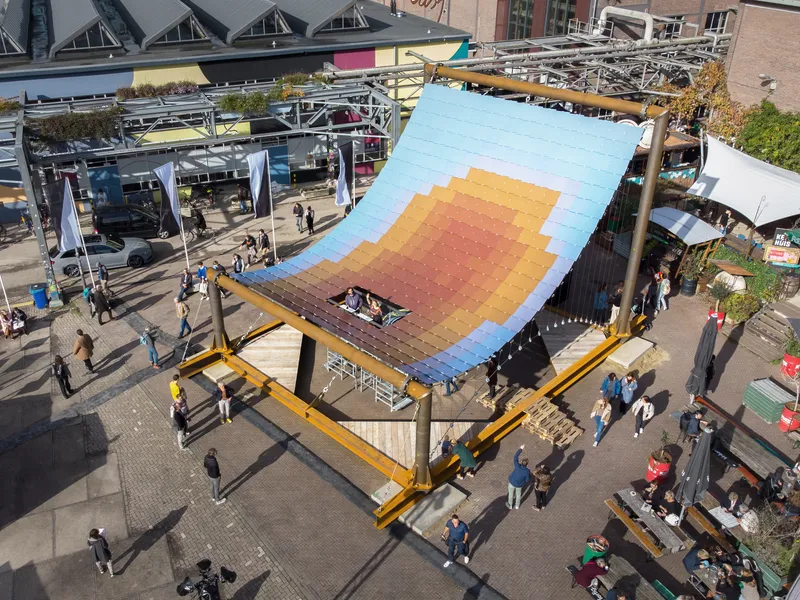Aerial view of tilted pavilion roof with gradient solar panels in blue, yellow, and orange over a public plaza