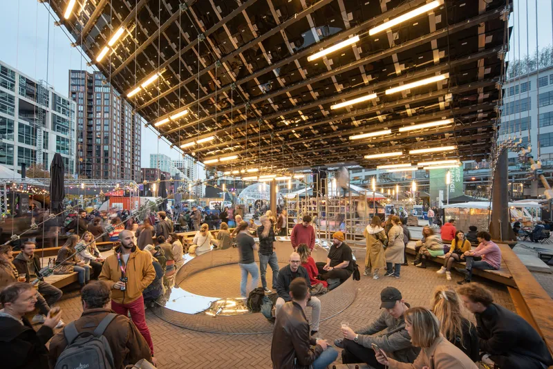 Underside of the timber grid ceiling with integrated lighting above a crowded public gathering space at dusk