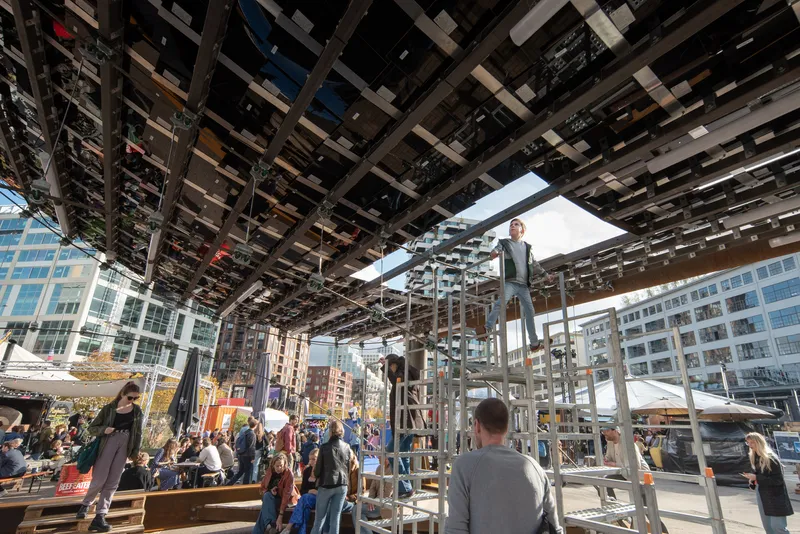 Looking up at a gridded steel canopy with rectangular openings as pedestrians gather beneath in daylight
