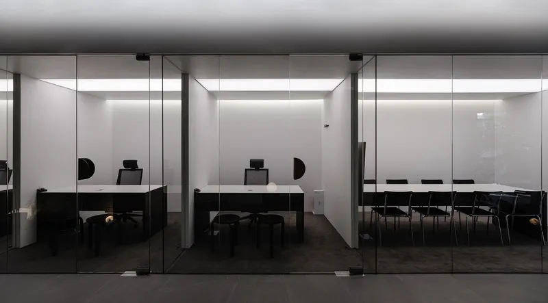 Glass-walled office spaces with black desks and chairs along a grey tiled corridor