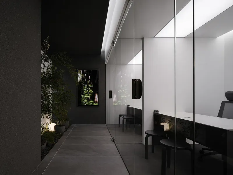 Covered outdoor corridor with dark paving and glazed wall reflecting interior workspace at night