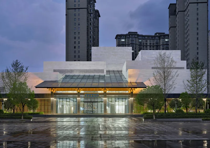 Glass entrance canopy illuminated at dusk with wet reflective paving in foreground and residential towers behind