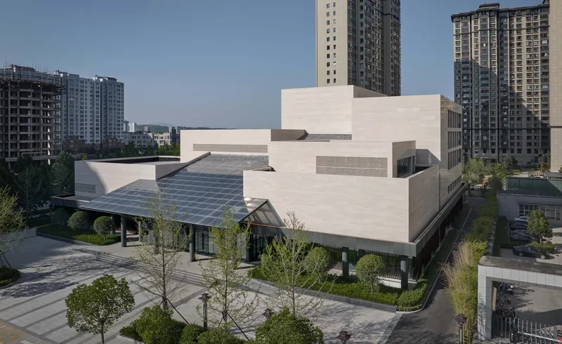 Tiered white stone volumes with solar panel canopy set among young trees and residential towers