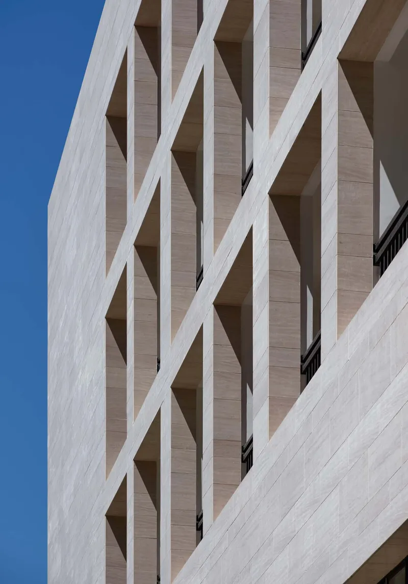 Detail of the travertine facade with diagonal white bracing elements forming an X-pattern against a clear blue sky